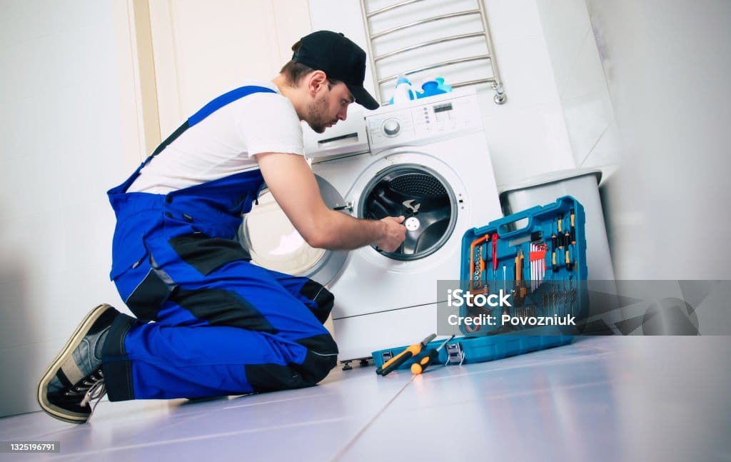 The young handsome repairman in worker suit with the professional tools box is fixing the washing machine in the bathroom
